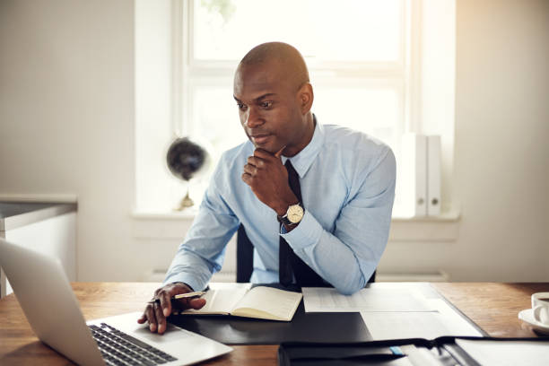 Young African business executive wearing a shirt and tie sitting at his desk in an office working online with a laptop